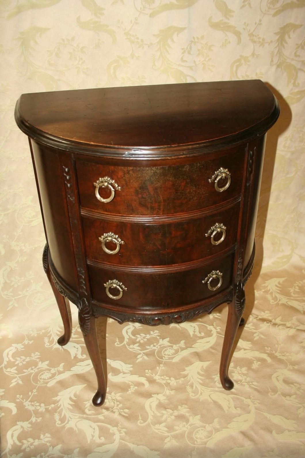 A demilune three-drawer nightstand in dark mahogany with brass ring pulls and cabriole legs